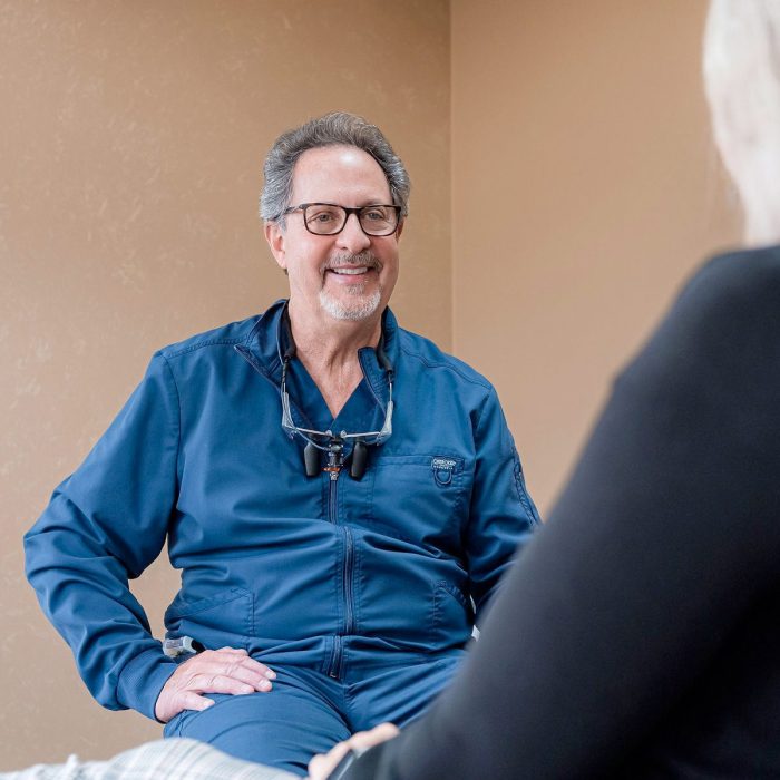 Dr. Lanik of Peak Dental Arts in Uniontown, OH wearing blue scrubs talking to patient with glasses around neck